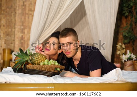Man and woman with fruits lie on bamboo bed with white linen in bedroom.