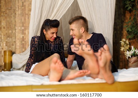 Talking barefoot man and woman smiles on bamboo bed with white linen in bedroom.