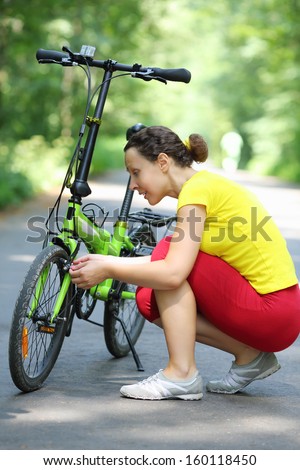 Young woman in sport clothes repairs bike in park at summer day.