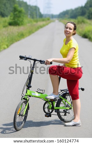Happy woman in sport clothes poses near bicycle on road at sunny day.