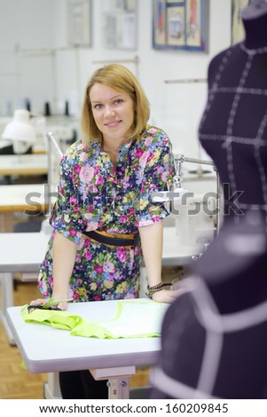 Female tailor stands near table with tailoring details and smiles.