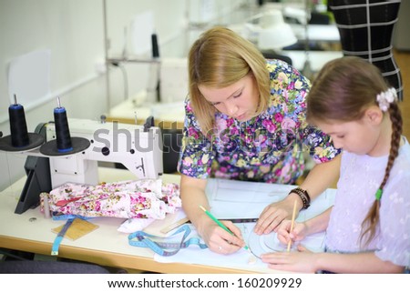 Female tailor stands near sewing machine and teaches student girl use of templates. Focus on woman.