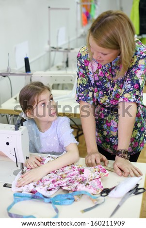 Pretty female tailor stands near table with sewing machine and talks with student girl. Focus on girl.