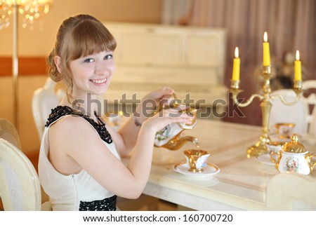 Pretty girl sits at classic white table with dishes and lighted candles and pours tea.