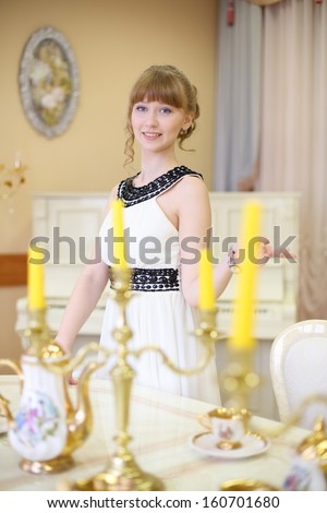 Beautiful smiling girl stands next to classic white table with set of dishes and candles.