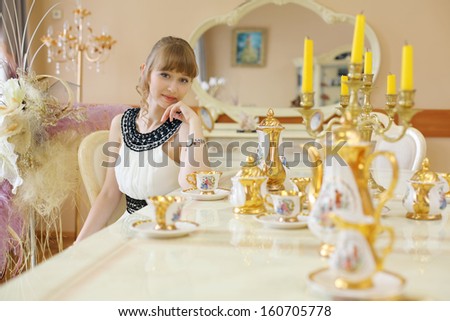 Beautiful girl in white dress sits at table with set of dishes and dreams.