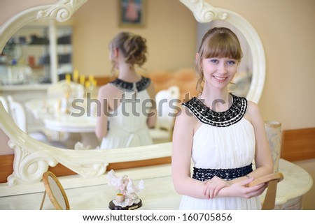 Beautiful smiling girl stands near mirror and holds frame with photo in light room.