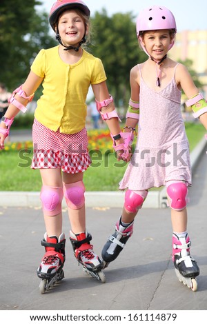 Two girls wearing helmets, elbow pads and knee pads ride on roller skates in the park