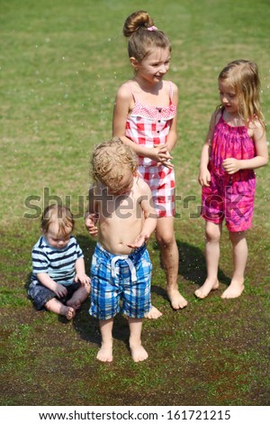 Four wet little funny children stand on grass among spray at summer day.