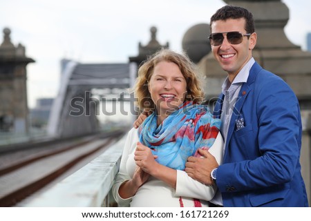 Pregnant woman in white and stylish man laugh near railroad bridge.