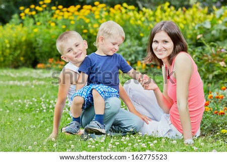 Young smiling woman sits on grass with two little sons on flower glade in park, youngest sits on brother knees