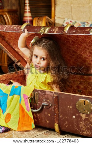 Little girl in yellow dress gets out from old big ragged fiber suitcase lying on floor in room