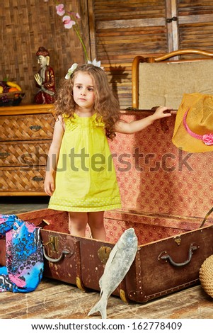Little girl in yellow dress stand in opened old big ragged fiber suitcase lying on floor in room