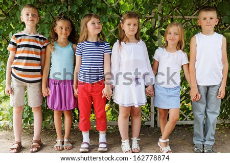 Five children friends stand holding hands in shadow under arch twined with green in summer park