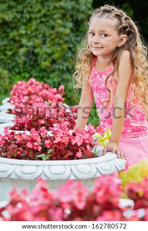 Little girl stands in summer park leaning on concrete flower pot full of flowers