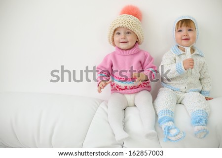 Two little kids in hats and knitted jackets sit on back of white sofa at home. Shallow depth of field.