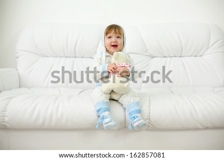 Little baby in hats and knitted suit with toy sits on white sofa at home. Shallow depth of field.