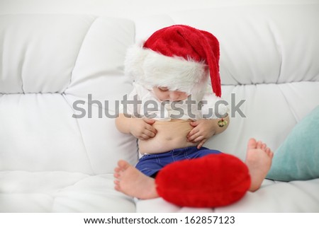 Cute little kid in red cap sits on white couch and looks at her belly at home. Shallow depth of field.