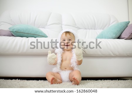 Barefoot baby in white soft pants, mittens and hat sits on carpet near white sofa and looks up at home. Shallow depth of field.
