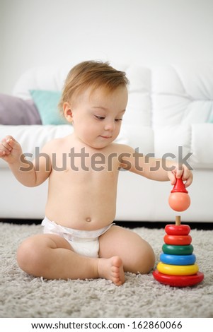 Cute little kid in nappy sits on carpet and plays with pyramid at home. Shallow depth of field.