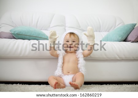Barefoot baby in white soft pants, mittens and hat sits on carpet near white sofa and smiles at home. Shallow depth of field.