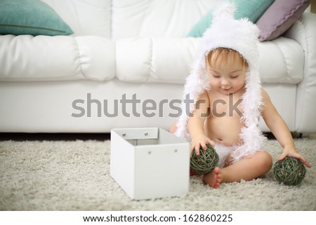 Pretty baby in white soft hat plays with balls and box near white sofa at home. Shallow depth of field.