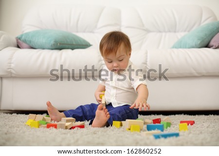 Barefoot baby sits on carpet and plays with wooden cubes near sofa. Shallow depth of field.