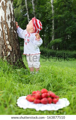 Girl in folk russian costume near birch tree, in the foreground is a plate with strawberries