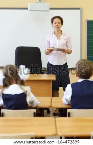 Teacher talks near chalkboard and pupils listen her at desks in classroom at school.