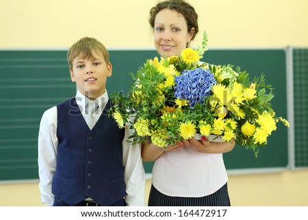 Happy teacher with big bunch of flowers and boy stand near chalkboard in classroom at school. Focus on boy.