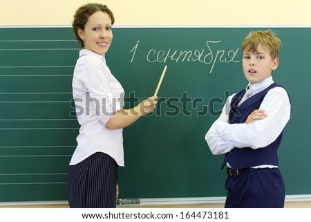 Boy and teacher with pointer stand near chalkboard with inscription September 1 in classroom at school. Focus on woman.