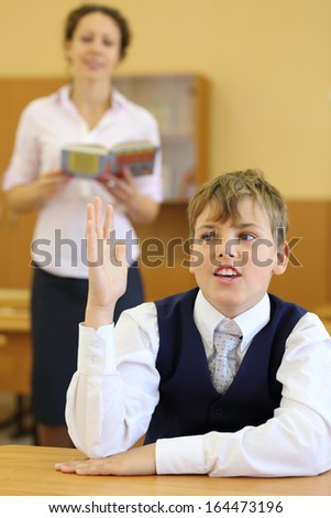 Teacher with book stands and boy sits at desk raised his hand to answer in classroom at school. Focus on boy.