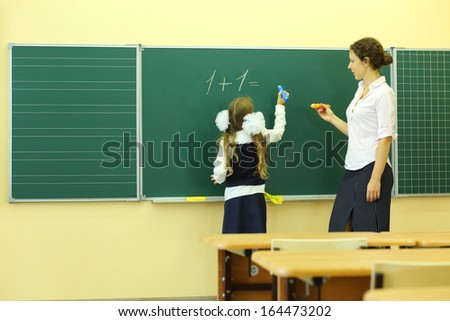 Girl and teacher near chalkboard solve simple math examples in classroom at school. Girl writes.