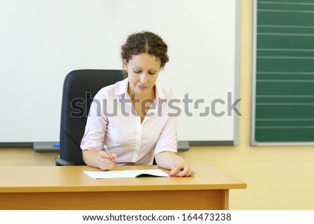 Young teacher in white shirt checks exercise book at desk in classroom.