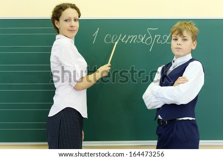 Boy and teacher stand near green chalkboard with inscription September 1 in classroom at school. Focus on woman.