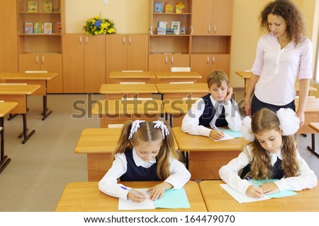 Two girls and boy sit at wooden school desks in classroom and teacher looks children work at school.