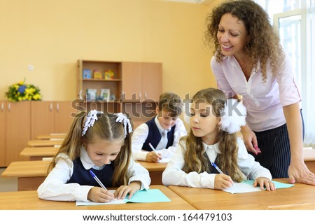 Two girls and boy write at school desks in classroom and happy teacher looks at exercise book at school.
