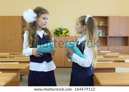 Two happy girls in uniform stand in classroom with exercise books and talk at school.
