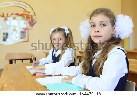 Two girls in school uniform sit at wooden school desk with exercise books in classroom at school. Focus on right girl.