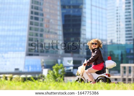 Little girl in leather jacket and sunglasses rides on toy motorbike on grass near skyscrapers.