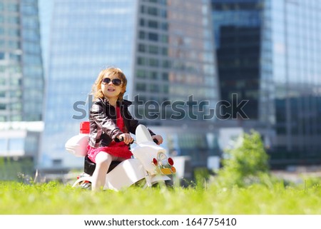 Little girl in leather jacket rides on toy motorbike on grass near skyscrapers.