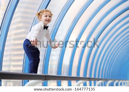 Little cute red hair girl balances on handrail in gallery near window.