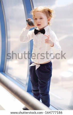 Little cute girl in white shirt talks by big black cell phone near window.