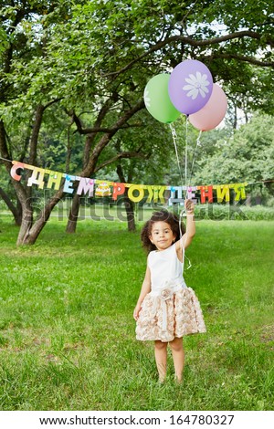 Smiling little girl stands in park, holding three air balloons in her raised hand, happy birthday sign behind her back