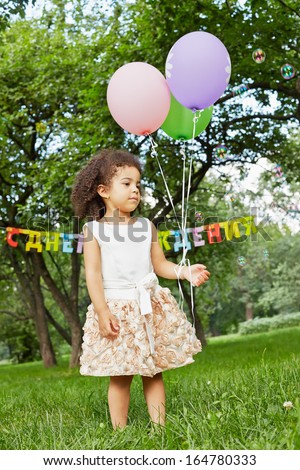 Little girl stands in park, holding three air balloons in her hand, soap bubbles fly around, happy birthday sign behind her back