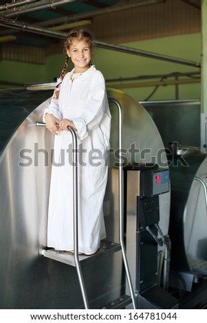Smiling little girl in white coat stands near big tank with milk at large farm.