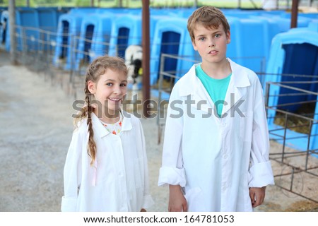 Pretty little girl and boy in white robes stands near blue pens for calves at cow farm. Focus on girl.