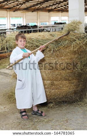 Smiling boy in white coat loads hay by big pitchfork at large cow farm.