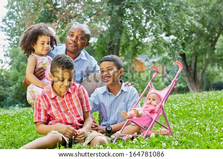 Father sits squatted and holds little daughter on his knee, two older sons sit nearby on grass