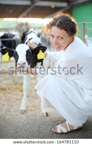 Woman in white robe caresses small calf and looks at camera in big cow farm.